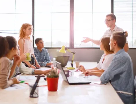 Группа из шести человек в светлом офисе с большими окнами, обсуждающая что-то за столом с ноутбуками и бумагами. Один человек стоит и оживленно говорит.